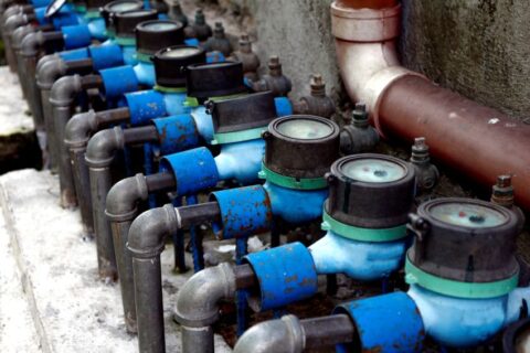Row of water meters connected to blue pipes along concrete wall